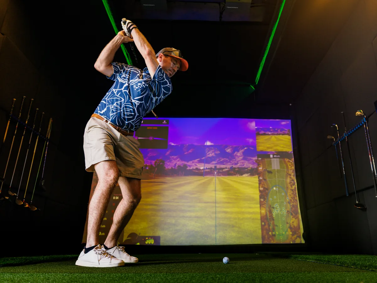 A golfer in an aloha shirt at the top of his swing inside the trailer, a fresh course loaded on the simulator screen