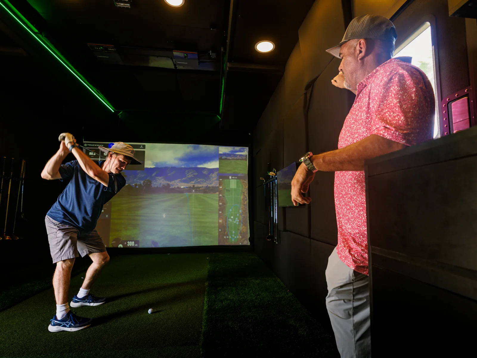 A golfer mid-swing inside the SwingLab trailer while Daren Kawika Martin watches from the counter, TrackMan tracking the shot on screen