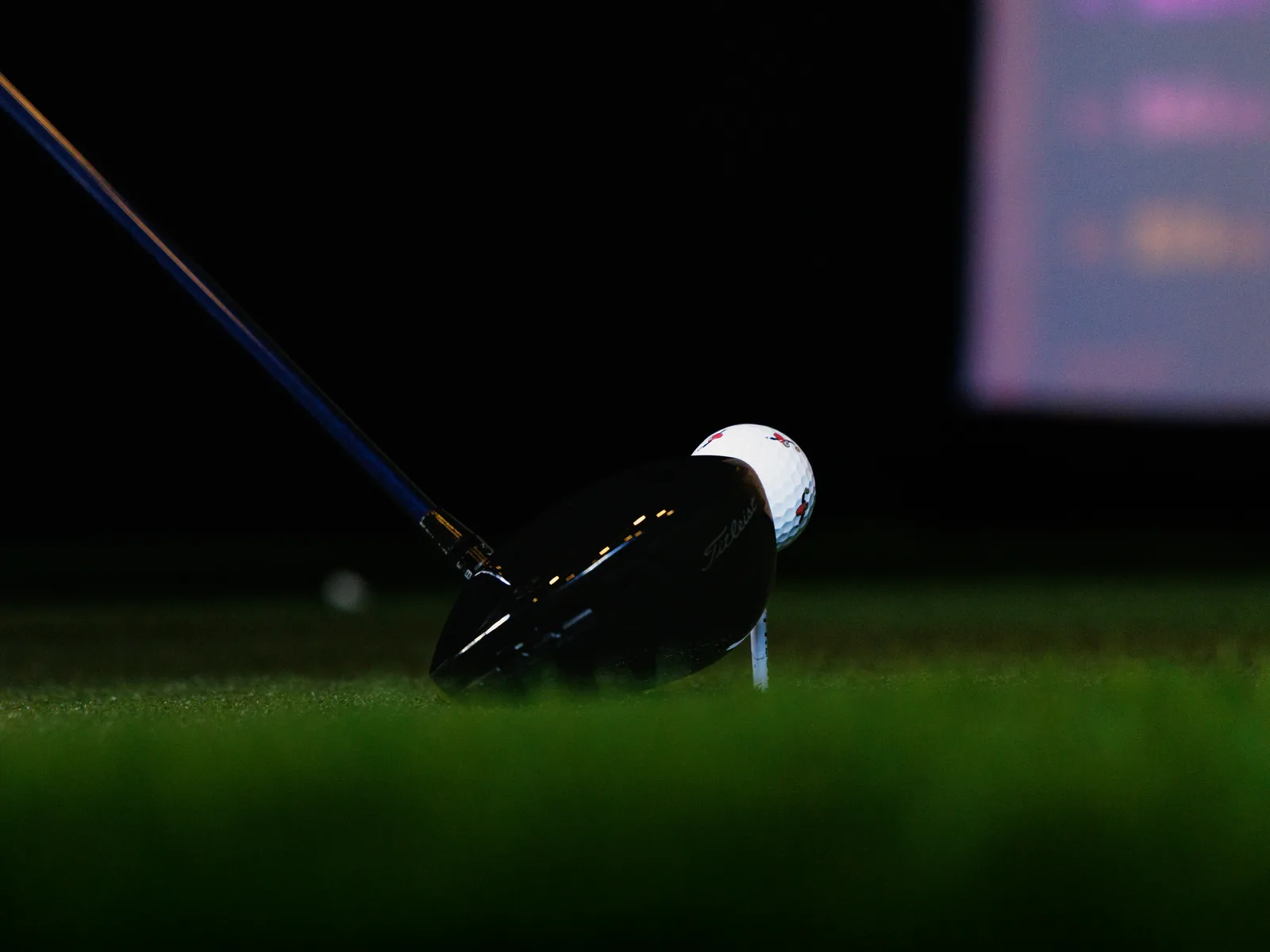 A driver head resting behind a ball on a tee on the turf inside the SwingLab trailer, TrackMan screen glowing behind it