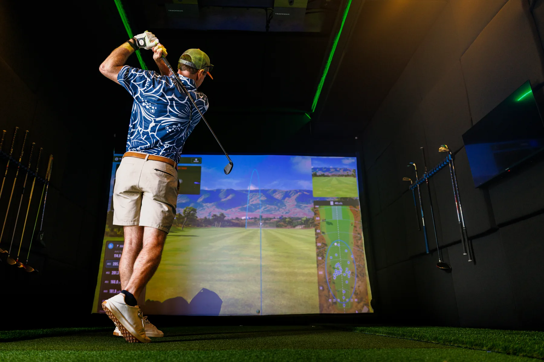 A golfer at the finish of his swing inside the SwingLab trailer, ball flight tracing across the TrackMan screen