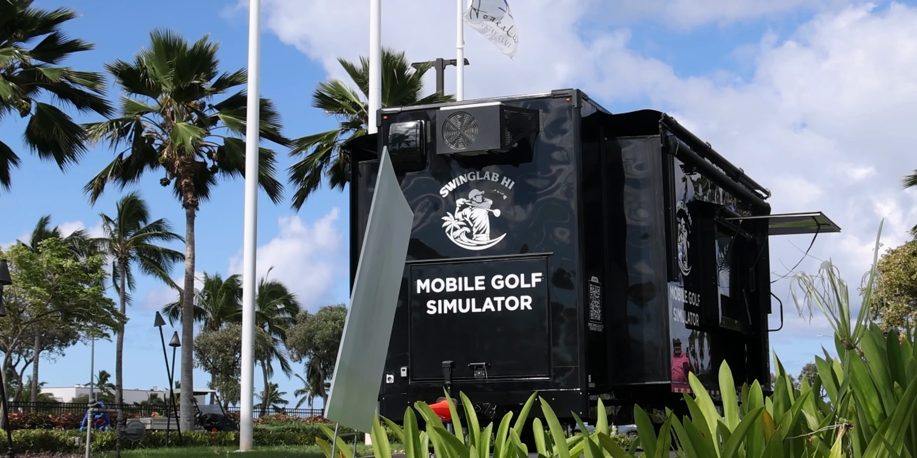 The SwingLab Hawaii mobile golf simulator trailer parked under palms at a club on Oahu, awnings out and a flag flying above it