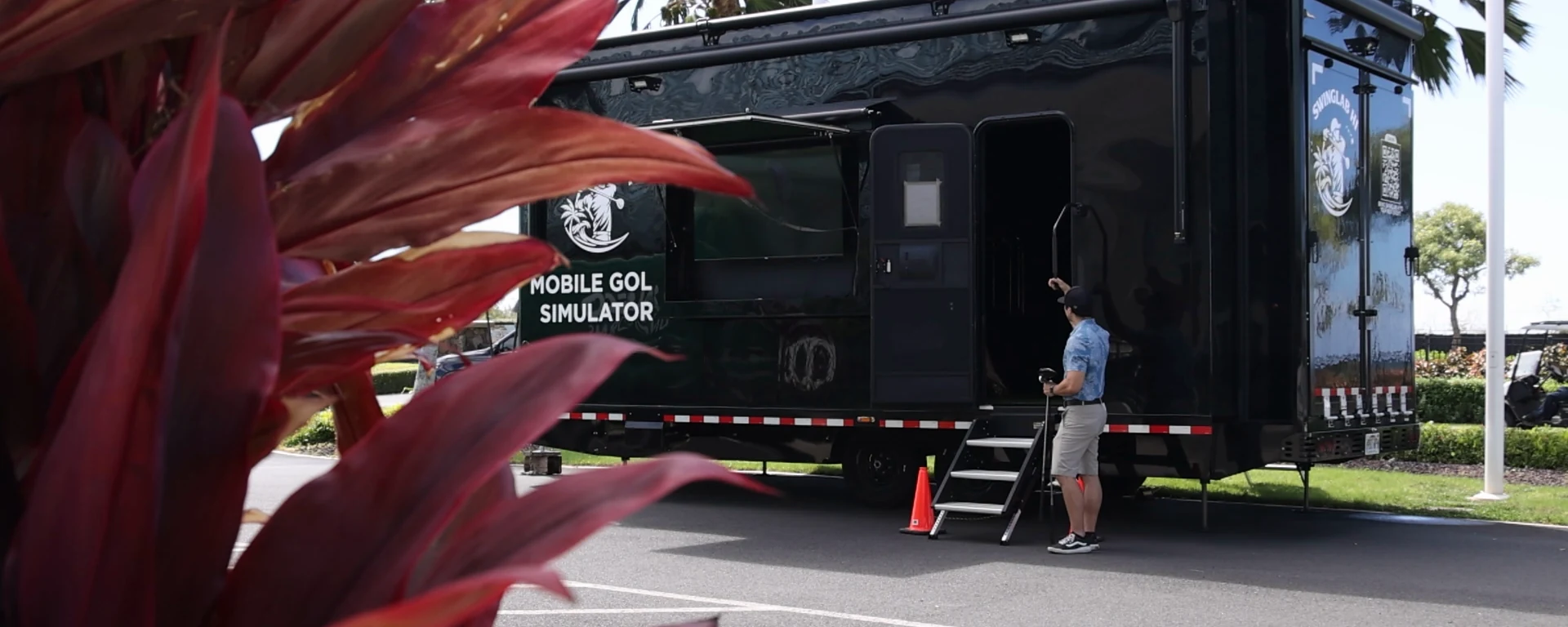 The SwingLab Hawaii mobile golf simulator trailer seen through red ti leaves, set up and open on Oahu